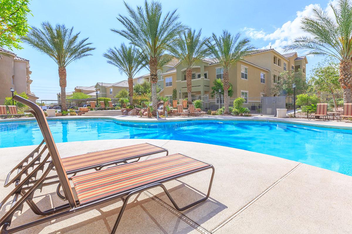A clear blue swimming pool surrounded by palm trees and lounge chairs, with a sunny sky and residential buildings in the background. The scene suggests a relaxing outdoor atmosphere ideal for leisure and enjoyment.