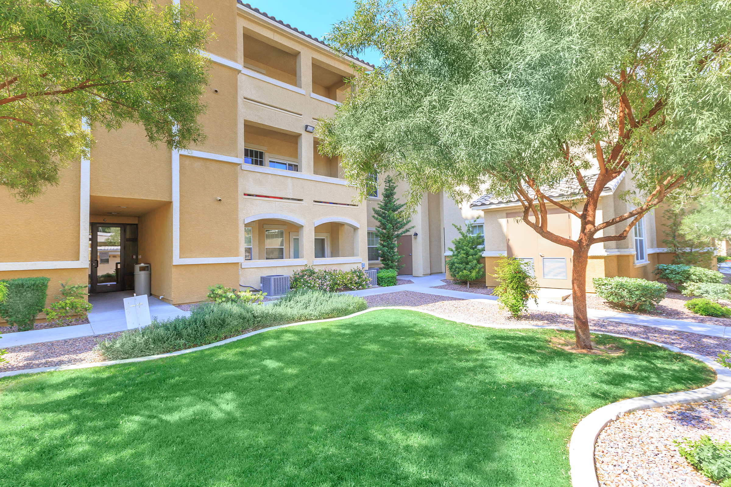 A sunny view of a landscaped residential area featuring green grass, shrubs, and trees. The image shows a modern building with multiple stories, a pathway, and a welcoming entrance. Bright blue skies enhance the inviting atmosphere of the community space.