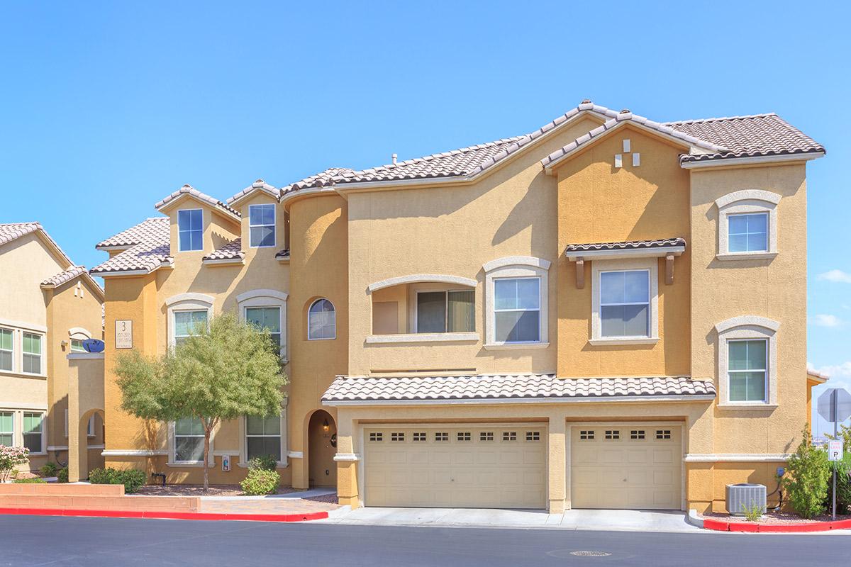 A two-story residential home with a light brown exterior and a tiled roof. The house features multiple windows, a front patio with greenery, and a three-car garage. The surrounding area has a clear blue sky and a curved red driveway, creating a welcoming suburban atmosphere.