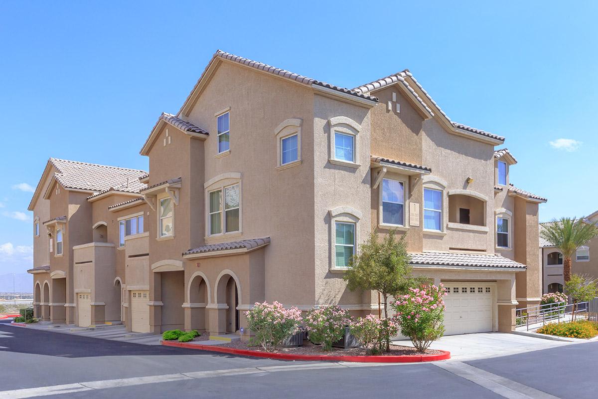 A corner view of a modern multi-unit residential building featuring beige stucco exteriors, tile roofs, and multiple windows. The scene includes well-maintained landscaping with shrubs and flowers, a paved driveway, and a clear blue sky in the background.