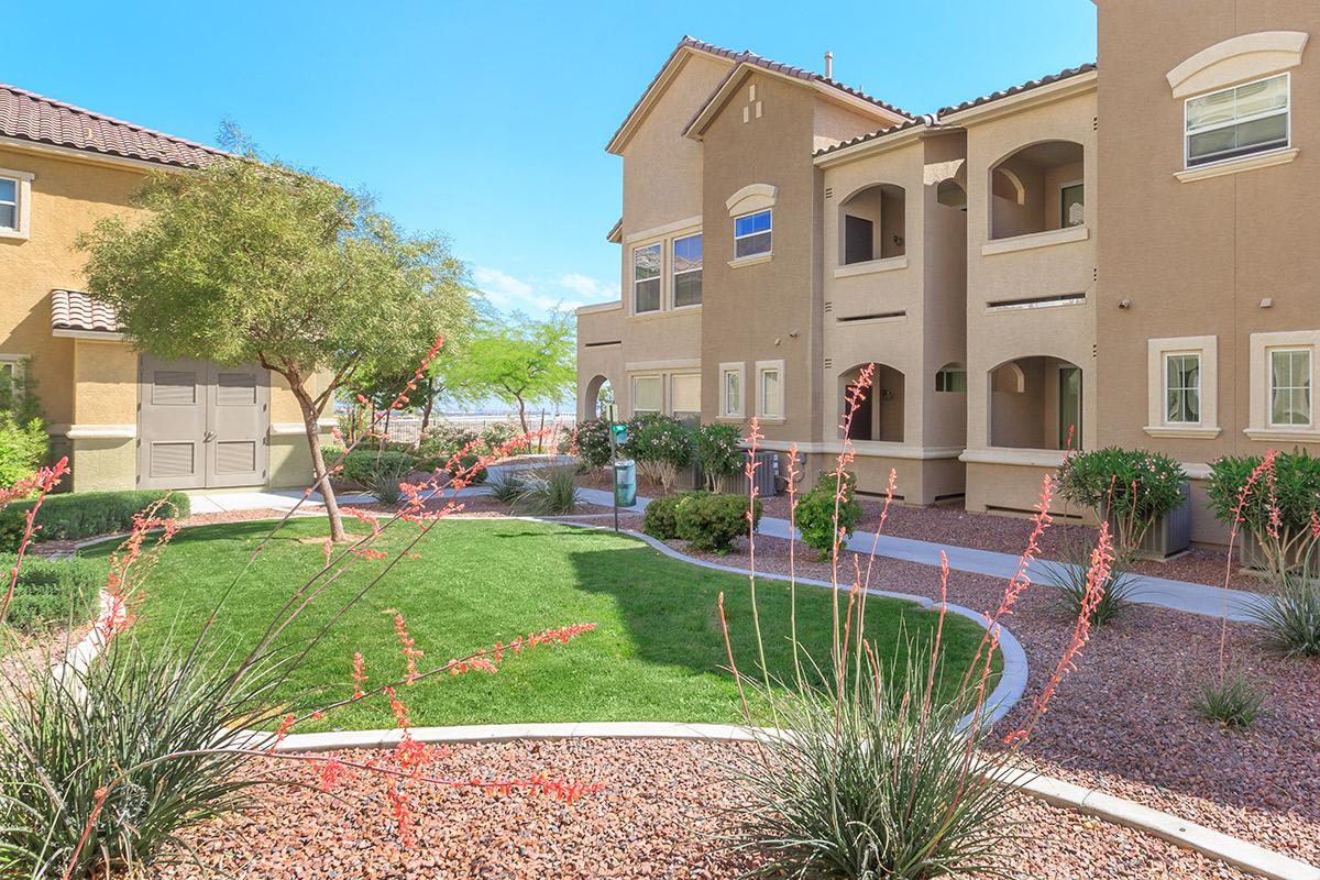 A landscaped courtyard featuring vibrant green grass and flowering plants, surrounded by elegant multi-story residential buildings. The clear blue sky and sunlight highlight the inviting atmosphere of the outdoor space.