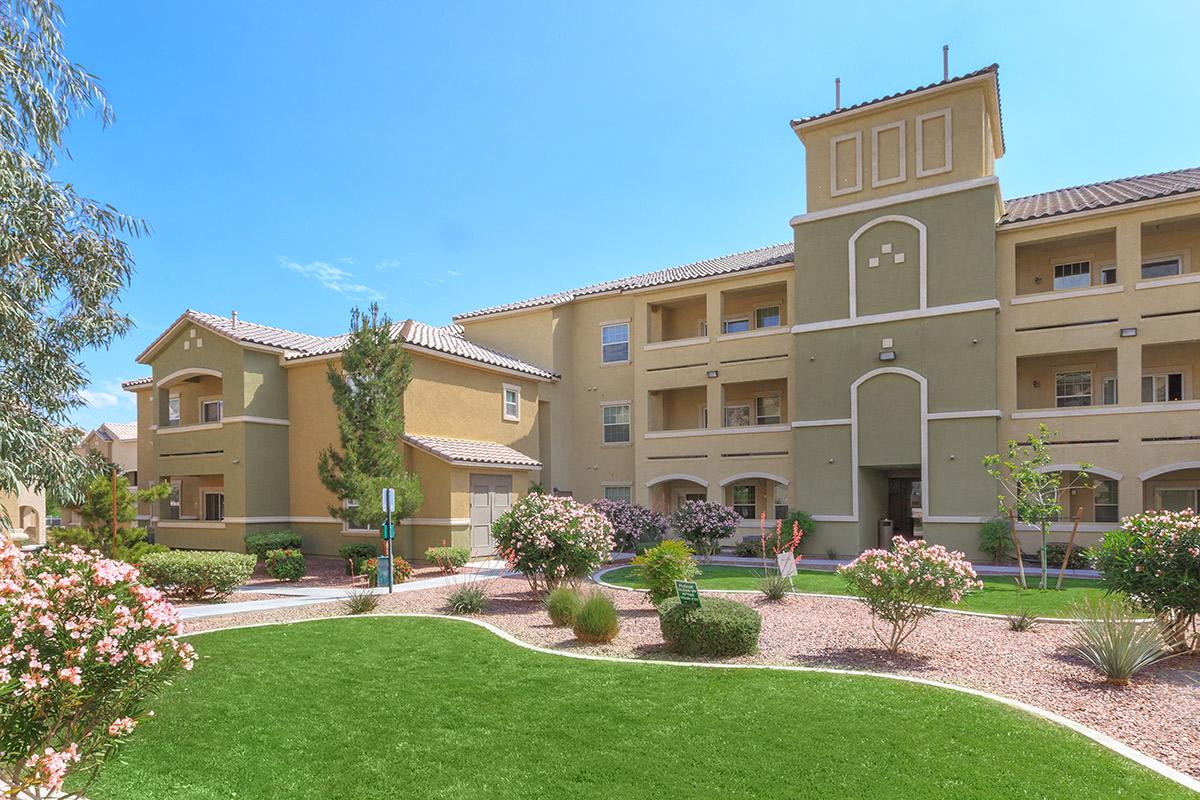 A multi-story apartment building with a beige and green exterior. It features several balconies, large windows, and a decorative tower. The surrounding area includes well-maintained landscaping with shrubs, flowers, and trees under a clear blue sky.