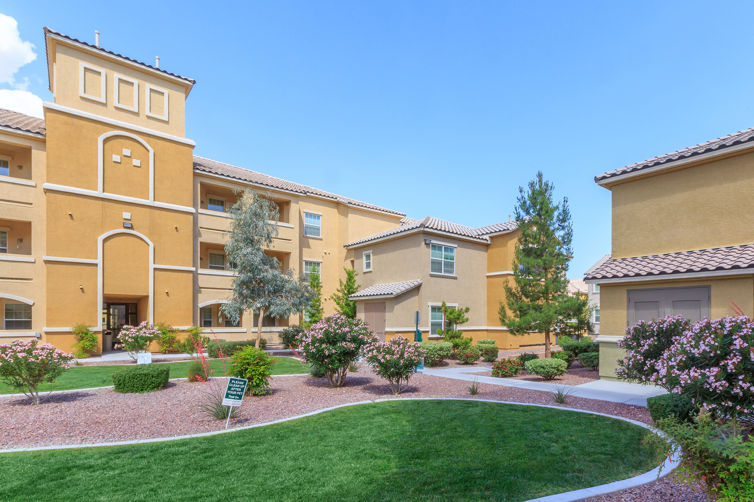 A modern apartment complex featuring beige and yellow buildings, landscaped green lawns, and flowering shrubs in a sunny outdoor setting. Blue skies with a few clouds are visible overhead, creating a bright and inviting atmosphere.