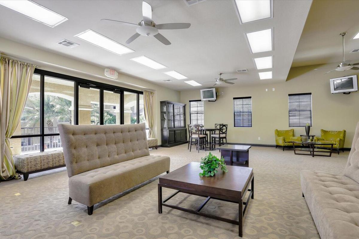 A spacious lounge area featuring a beige sectional sofa, a wooden coffee table with a small plant on it, and casual seating arrangements. Large windows provide natural light, and ceiling fans help with ventilation. The room has a neutral color scheme and modern decor, making it inviting and comfortable for gatherings.
