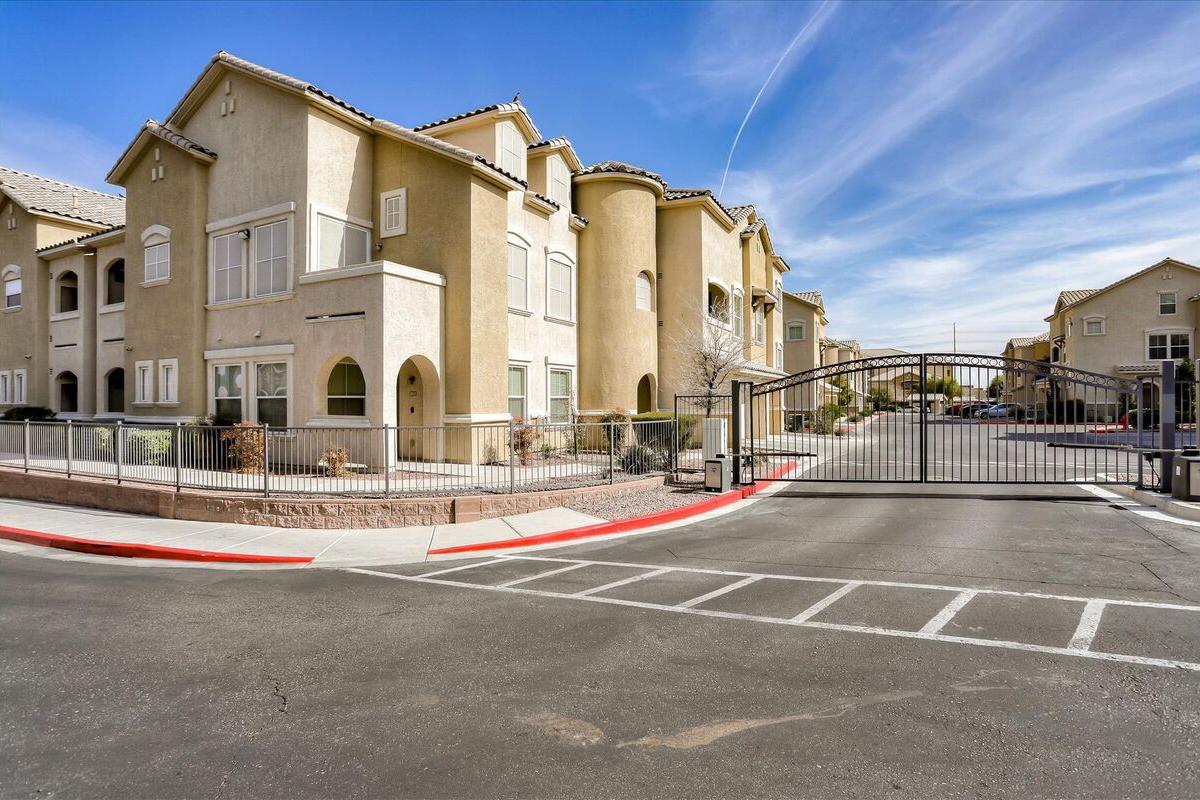 A row of beige apartment buildings with multiple windows and balconies, set against a clear blue sky. A gated entrance leads into the residential complex, with a red curb and parking area visible in the foreground. The scene conveys a quiet suburban atmosphere.