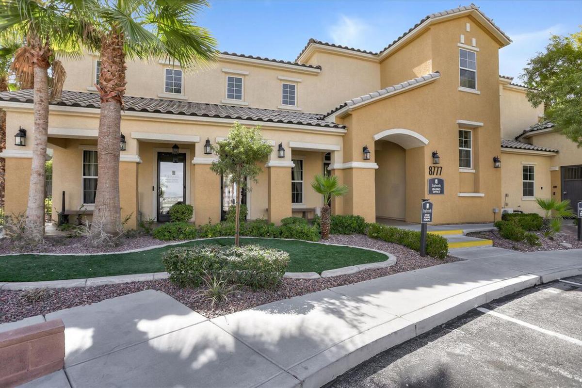 A yellow two-story building with multiple windows and a welcoming entrance, flanked by palm trees and neatly landscaped greenery. A small sign near the entrance displays the building number, and a paved parking area is visible in front. The sky is clear and blue.