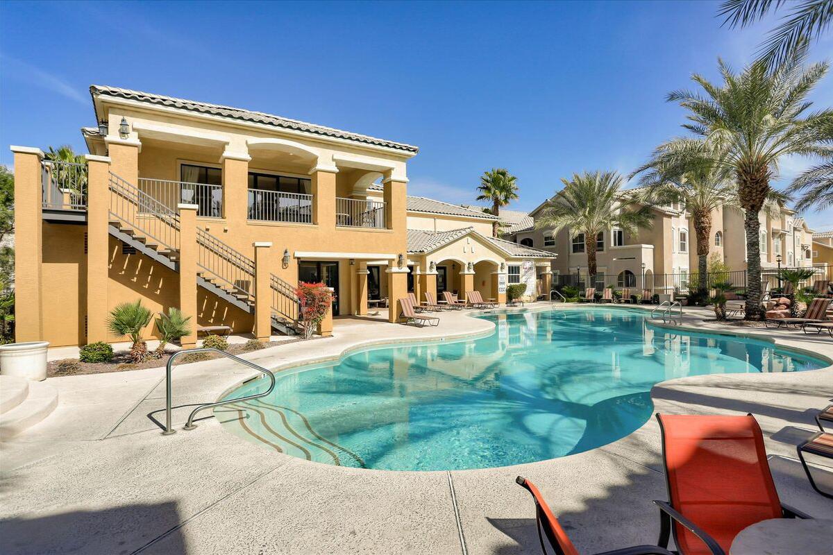 A spacious poolside area featuring a clear blue swimming pool surrounded by lounge chairs. The area is lined with palm trees and has a stylish building with a staircase and balconies overlooking the pool, creating a relaxing resort-like atmosphere on a sunny day.