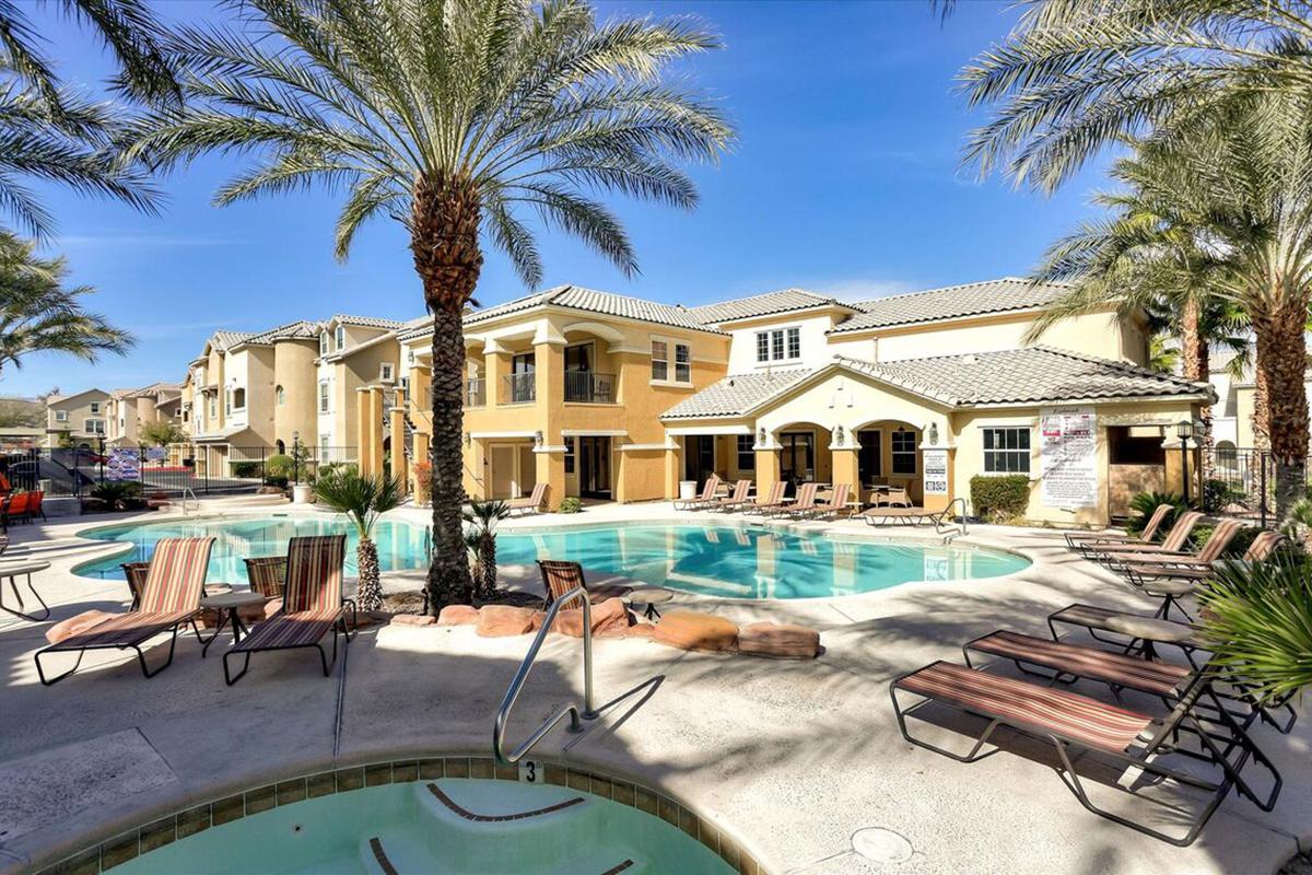 A sunny outdoor pool area surrounded by palm trees, with lounge chairs and a hot tub. In the background, several multi-story buildings can be seen, creating a relaxed atmosphere. Clear blue skies enhance the inviting setting.