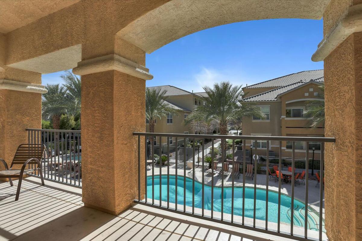 View from a balcony overlooking a landscaped courtyard with swimming pools and lounging chairs. Palm trees and buildings are visible in the background under a clear blue sky. The scene suggests a relaxing outdoor space perfect for leisure activities.