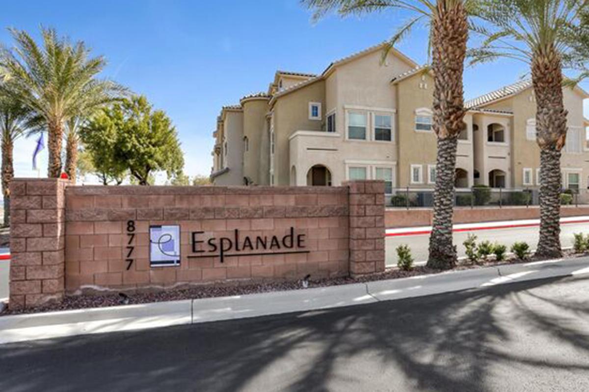 Sign for "Esplanade" at 8777, displayed on a stone wall. The background features a residential building with several palm trees and clear blue skies. The area is well-maintained, with landscaping visible along the pathway.