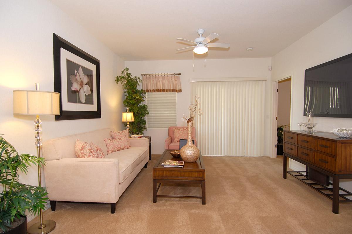 A cozy living room featuring a light-colored sofa with decorative pillows, a wooden coffee table, and a console table against the wall. A large mirror and a framed floral artwork enhance the decor, while a ceiling fan and lamps provide lighting. Potted plants add a touch of greenery to the space.