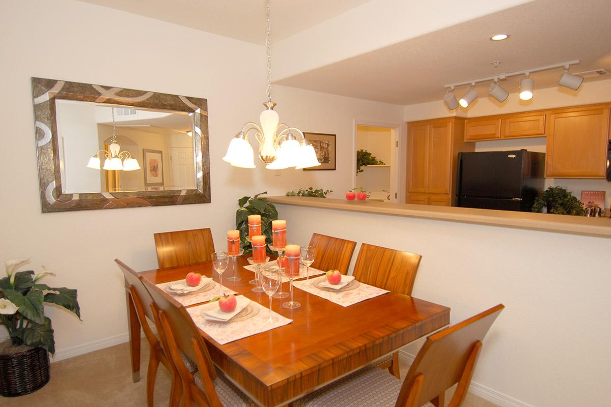 Modern dining area featuring a wooden table set for four, complete with decorative plates and candles. A pendant light hangs above the table. The walls are painted white, and there's a large mirror reflecting the space. In the background, a kitchen with wooden cabinets is visible. A potted plant is positioned in the corner.