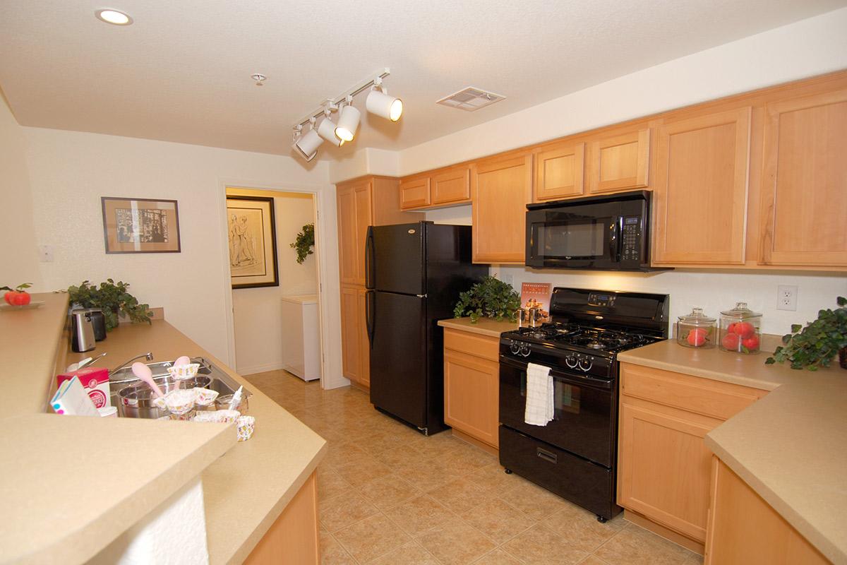 A modern kitchen featuring light wooden cabinets, a black refrigerator, and a black gas stove. The countertop is beige, with decorative green plants and jars in the background. It has track lighting and an open layout that leads to another room. The overall design is warm and inviting.