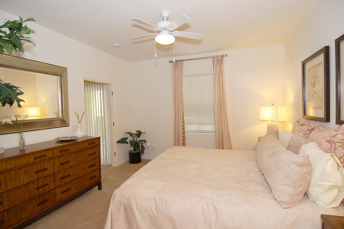 A cozy bedroom featuring a large bed with light bedding, a wooden dresser, and bedside lamps. There is a full-length mirror on the wall, a ceiling fan, and a window with sheer curtains. Decorative plants add a touch of greenery to the space, creating a warm and inviting atmosphere.