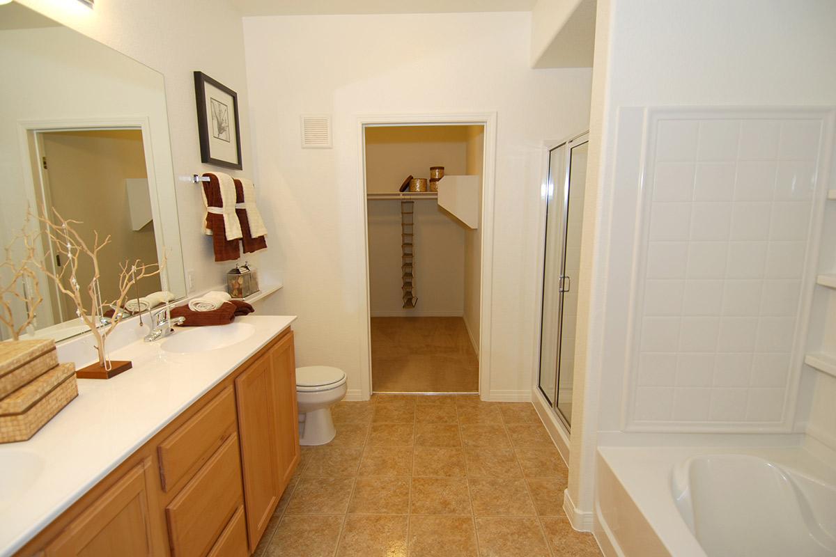 A spacious bathroom featuring a double sink vanity with wooden cabinetry, a shower enclosure, a toilet, and a bathtub. The room has beige tiled flooring and a large mirror. A doorway leads into a walk-in closet, and decorative towels hang on a rack. Natural light brightens the space, creating a warm atmosphere.