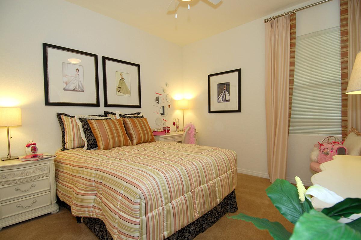 A cozy bedroom featuring a neatly made striped bed with decorative pillows. The walls showcase framed fashion illustrations, and there are soft lighting fixtures beside the bed. A small dressing table and a plush chair add to the inviting atmosphere, complemented by light-colored curtains.