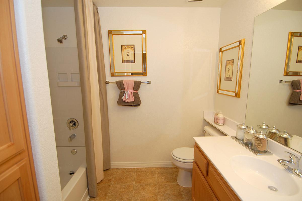 A clean and modern bathroom featuring a beige tile floor, a bathtub with a shower curtain, a toilet, and a double sink vanity. The walls are painted white, and there are decorative elements like framed art, towels, and a set of glass containers on the counter.