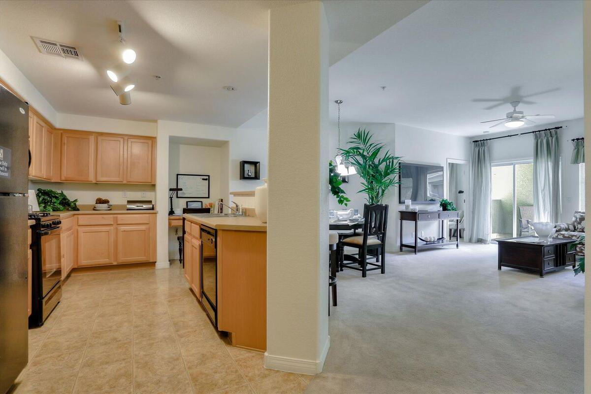 A spacious, modern open-concept living area featuring a kitchen with wooden cabinetry, stainless steel appliances, and a dining space. A comfortable seating area is visible, adorned with plants and natural light from large windows, creating a welcoming atmosphere.