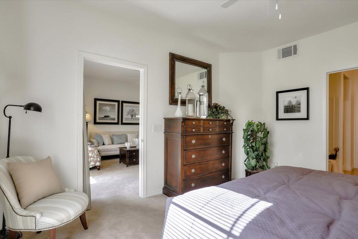 A cozy bedroom featuring a wooden dresser, a soft chair, and a large mirror. Sunlight streams in, casting shadows on the carpeting. The open doorway leads to a living area with a couch and decorative artwork on the walls, creating a warm and inviting atmosphere.