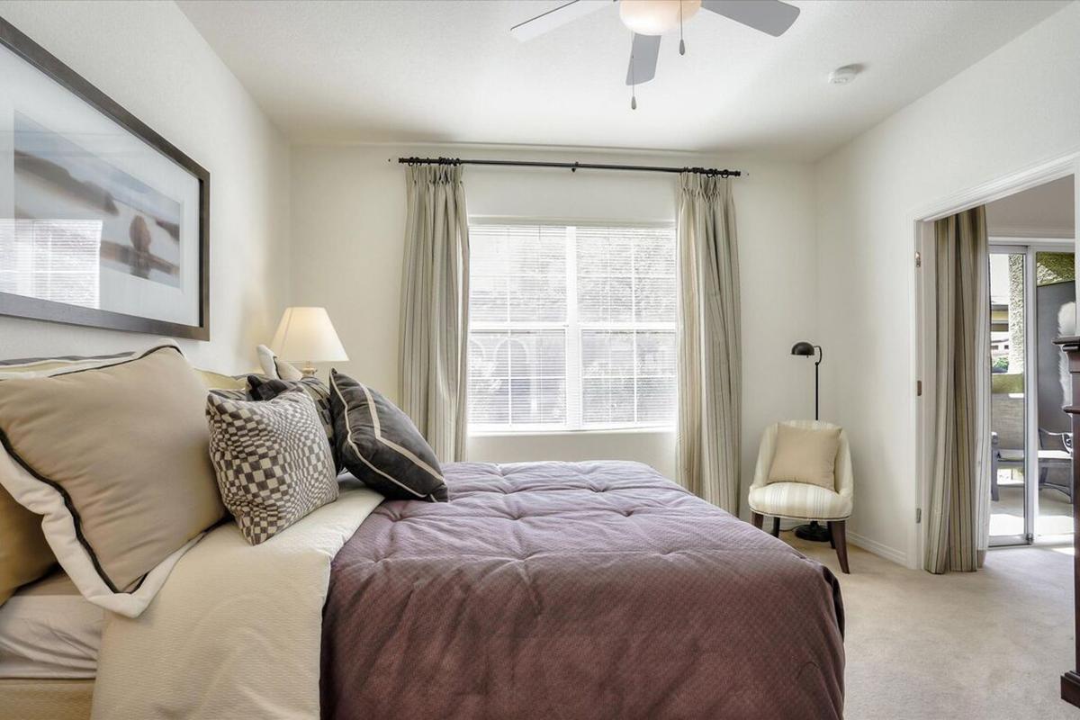 A cozy bedroom featuring a queen-sized bed with decorative pillows, a bedside lamp, and a ceiling fan. Light-colored walls and curtains enhance the bright atmosphere, while a window allows natural light in. A chair in the corner and an adjacent doorway lead to an outdoor area.