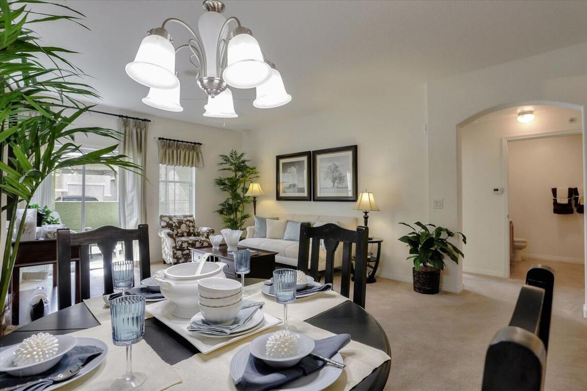 A cozy dining area set for a meal, featuring a dark wooden table with blue glasses, white dishes, and decorative items. A large light fixture hangs overhead, and the background includes a living space with comfortable seating, plants, and wall art. A doorway leads to another room.