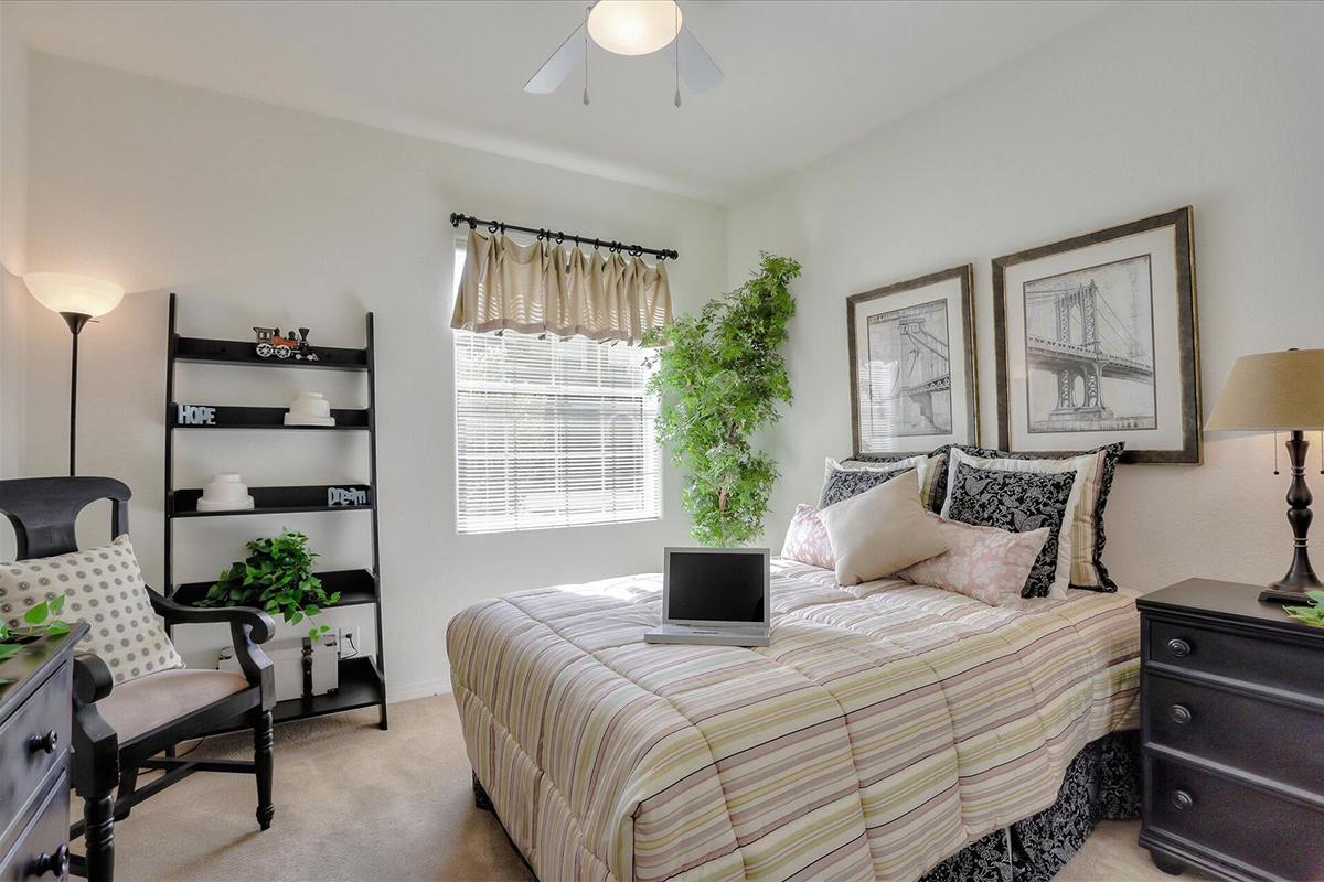 Cozy bedroom with a neatly made bed featuring striped bedding, a laptop on the bed, and decorative pillows. A window with sheer curtains allows natural light, complemented by two framed artworks on the wall. A small bookshelf and a floor lamp add a warm ambiance, while a potted plant provides a touch of greenery.