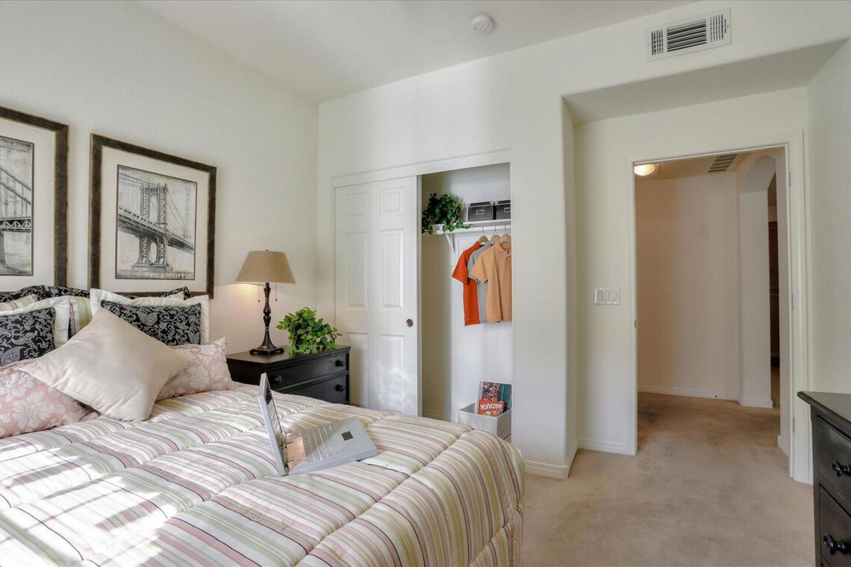 Cozy bedroom featuring a bed with decorative pillows, a bedside lamp, and framed artwork on the walls. A closet with hanging clothes is visible, along with light-colored walls and carpeted flooring. A doorway leads to a hallway, creating a bright and inviting atmosphere.