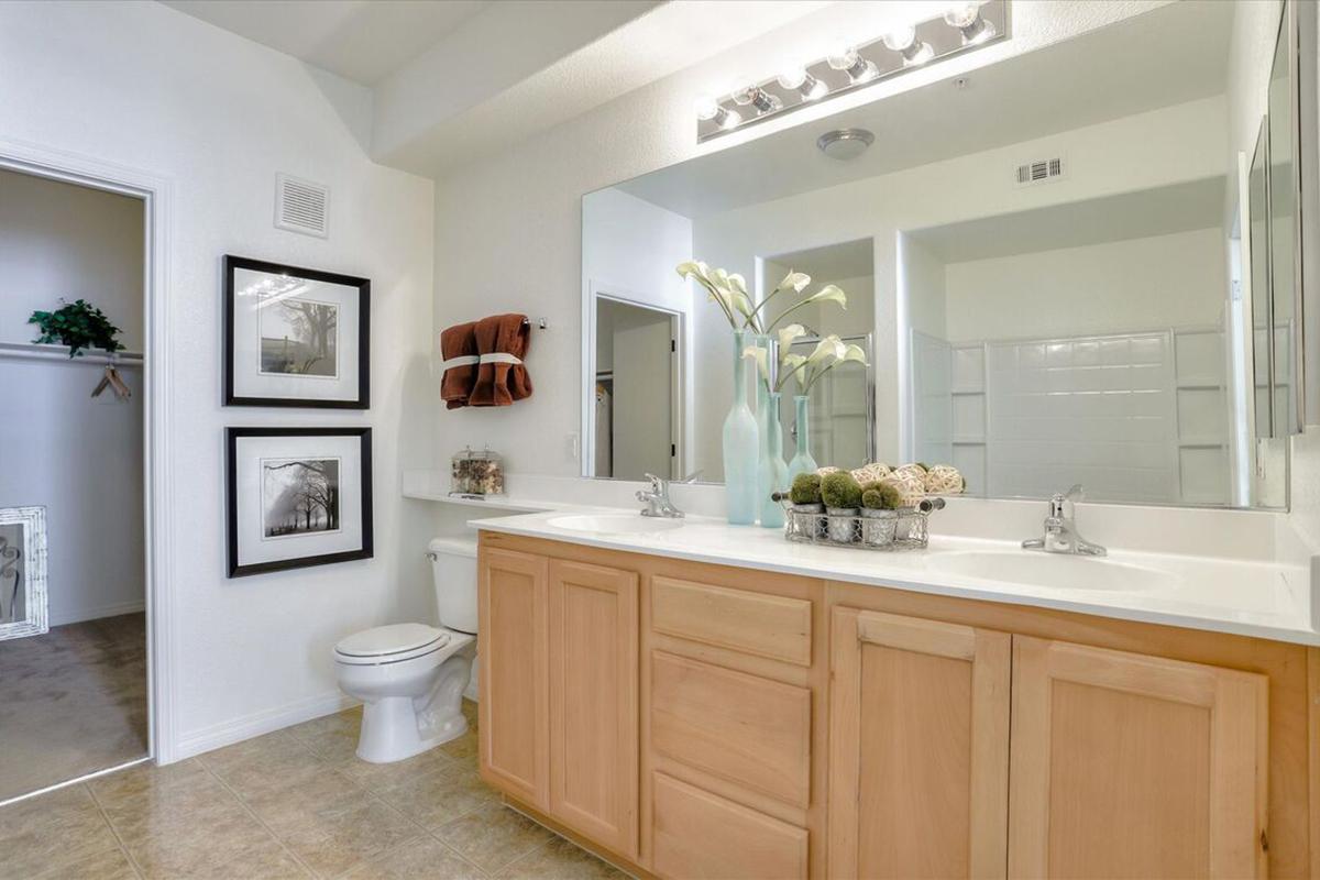 A modern bathroom featuring a light-colored vanity with a white countertop, framed artwork on the walls, and a mirror above the sink. Decorative items, including a vase with flowers and a bowl of stones, are placed on the countertop. The space is bright and airy, with a clean design and neutral color palette.