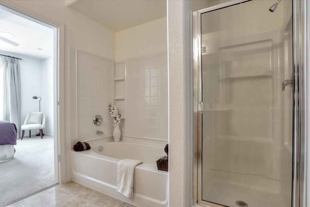 A modern bathroom featuring a bathtub on the left with towels and decorative items, and a separate glass shower on the right. The walls are painted white, and there's a door leading to another room in the background with partial visibility of a chair and window. Natural light brightens the space.