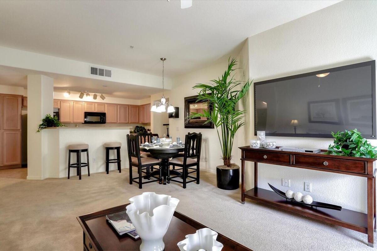 A modern living room and dining area featuring a dark wood table set for four, a large flat-screen TV mounted on the wall, and light-colored walls. A potted plant adds greenery, while a coffee table displays decorative items. The kitchen area with barstools is partially visible in the background.