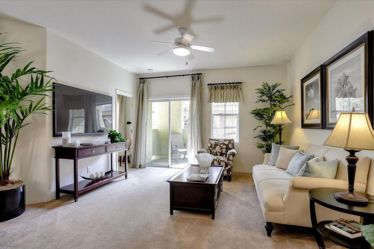 A cozy living room featuring a light-colored sofa, a wooden coffee table, and decorative plants. There's a flat-screen TV mounted on the wall, a stylish armchair, a lamp, and large windows with sheer curtains letting in natural light. A sliding door leads to a balcony area. The decor is warm and inviting.