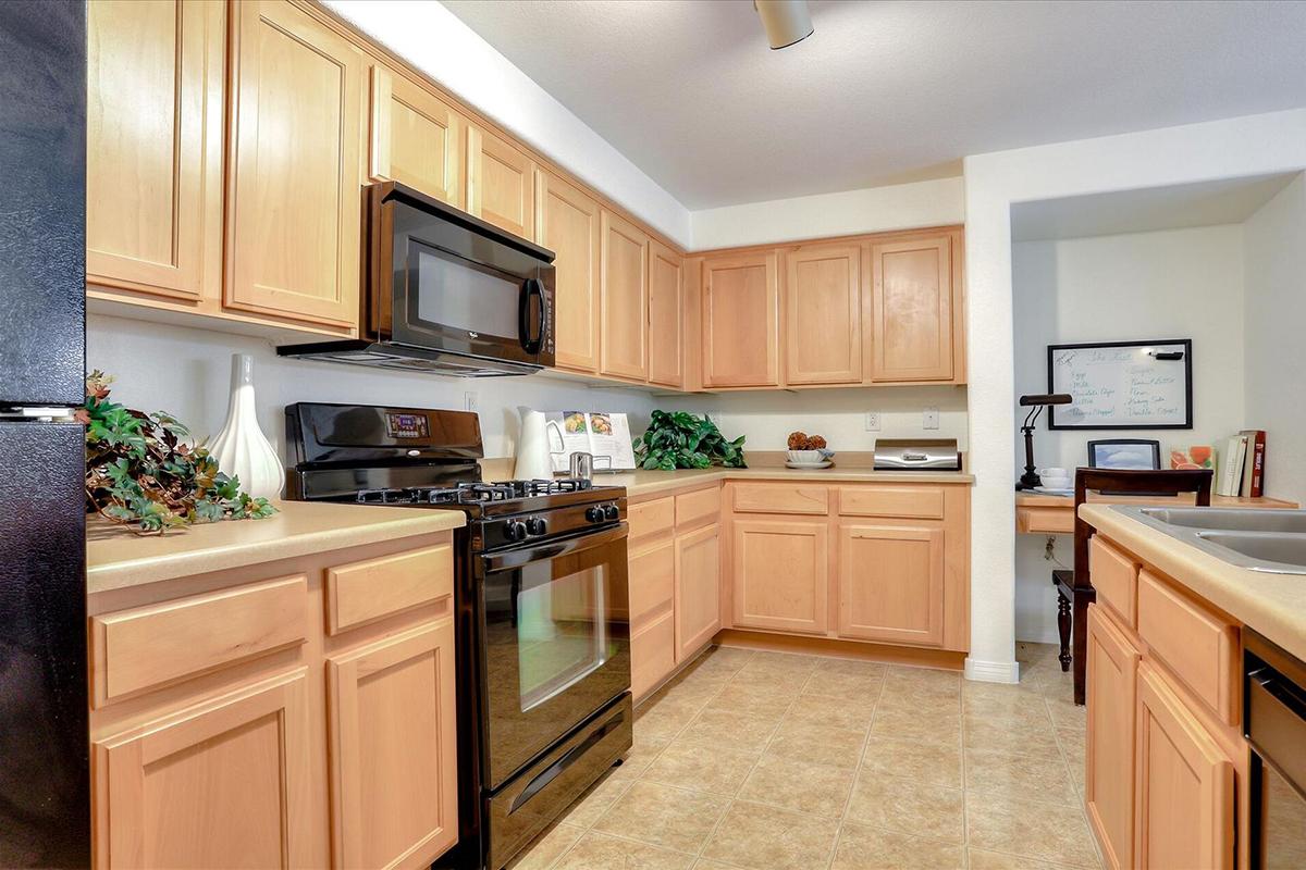 A modern kitchen with wooden cabinetry, a black stove, microwave, and sink. Counter space is used for a few kitchen items, with decorative plants adding a touch of greenery. The room has neutral-colored walls and tile flooring, creating a warm and inviting atmosphere.