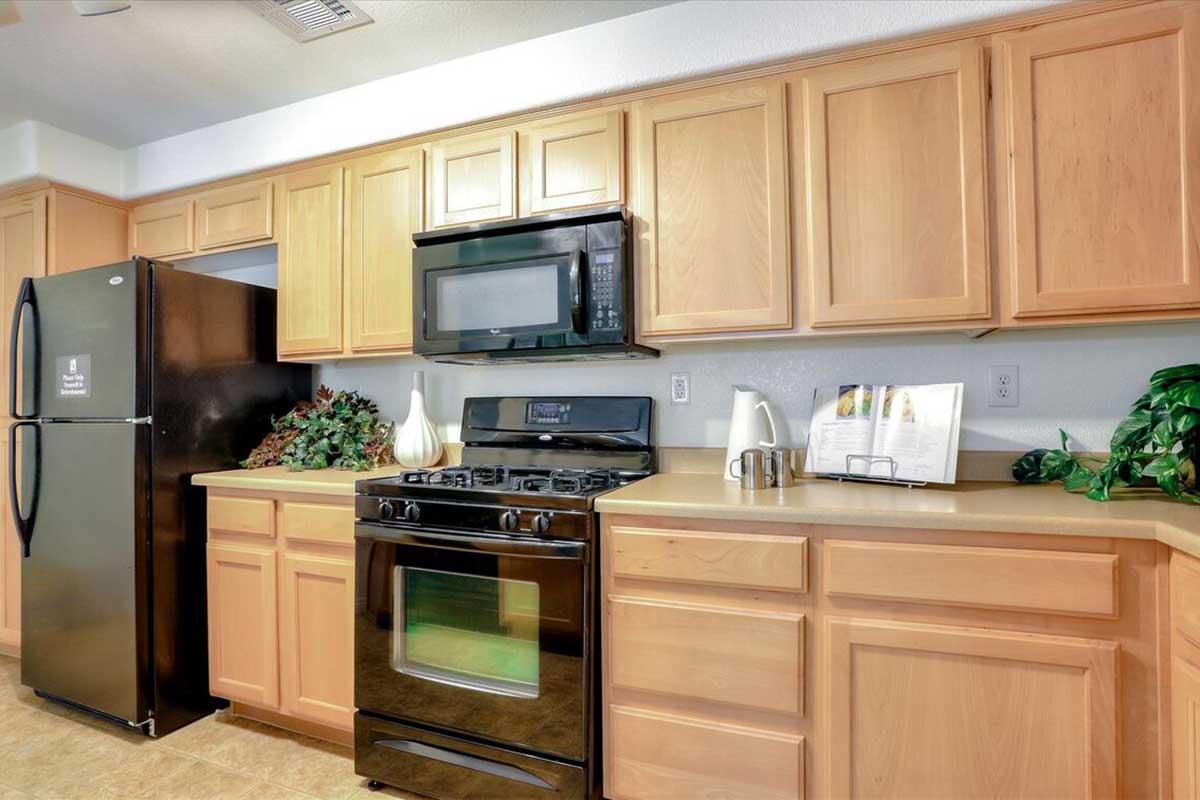 A modern kitchen featuring light wooden cabinets, a black refrigerator, and a black gas stove. There is a microwave above the stove and a small countertop with a kettle and decorative plants. A framed picture is displayed on the counter, complementing the overall design.