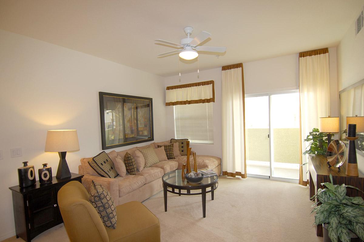 A bright, modern living room featuring a beige sectional sofa with decorative pillows, a round glass coffee table, and a black entertainment unit. A lamp on a side table and a ceiling fan add to the decor. Large windows with light curtains provide natural light, and a balcony door is visible.