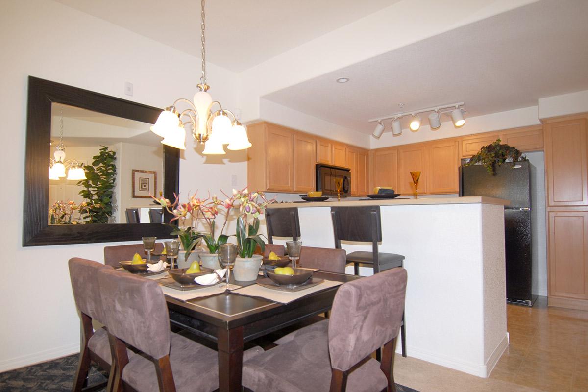 Dining area in a modern kitchen featuring a wooden table set with plates and decorative flowers. In the background, there's a kitchen bar with stools, light cabinetry, and a black fridge. A large mirror reflects the space, enhancing the bright and inviting atmosphere.
