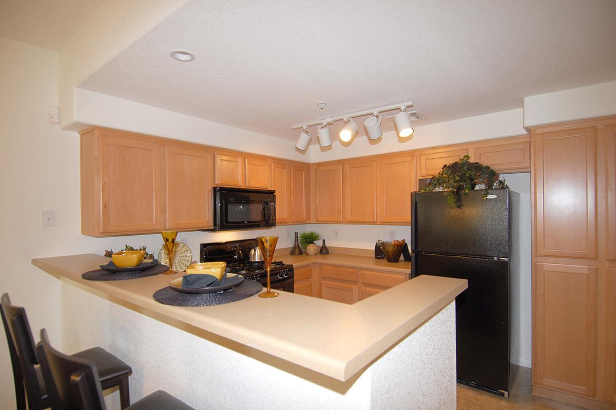 A modern kitchen featuring light wooden cabinetry, a black refrigerator, and a black microwave. The counter has two place settings with decorative plates and glasses. Recessed lighting illuminates the space, creating a warm and inviting atmosphere. The flooring appears to be a light tile.