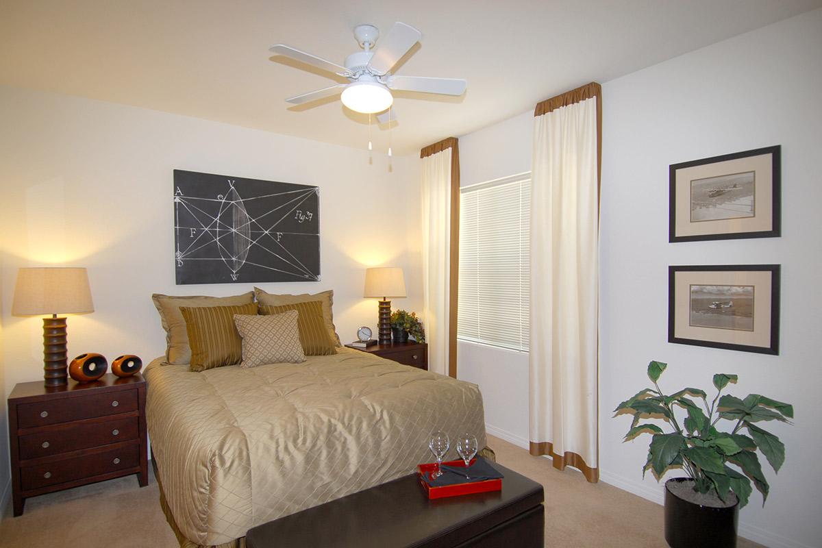 Cozy bedroom featuring a queen-sized bed with neutral bedding and decorative pillows, a ceiling fan, two bedside lamps, framed art on the walls, a window with light curtains, and a potted plant in the corner. A small table with two wine glasses sits at the foot of the bed.