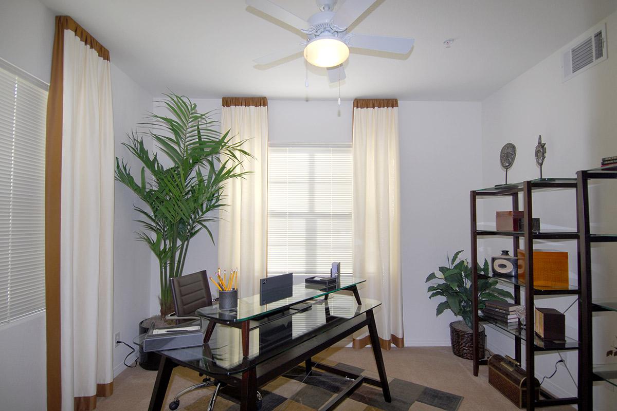 A modern office space featuring a sleek glass desk with an ergonomic chair, surrounded by large windows with sheer curtains. There are decorative plants in the corners and a bookshelf filled with various items, creating a bright and inviting work environment.