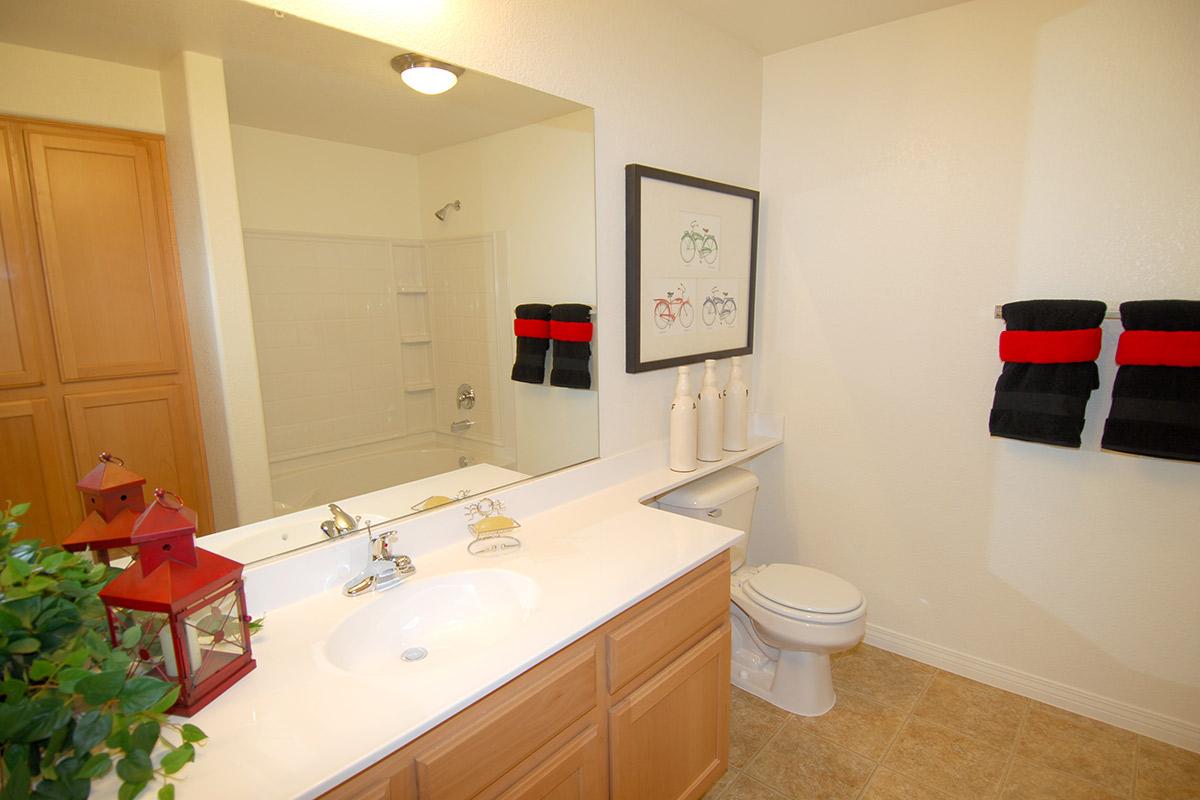 A well-lit bathroom featuring a large mirror, a white countertop with sinks, and wooden cabinetry. Decorative items include a red and black towel set, a small plant, and a red lantern. The space includes a shower area with white tiles and neutral-colored walls, creating a clean and modern look.
