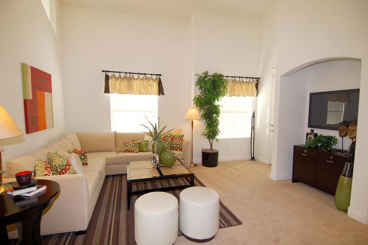 A bright, modern living room featuring a beige sofa with colorful cushions, two white ottomans, a coffee table with plants, and a floor lamp. There are large windows with curtains, a television mounted on the wall, and decorative plants. The space is elegantly styled with a light color palette.