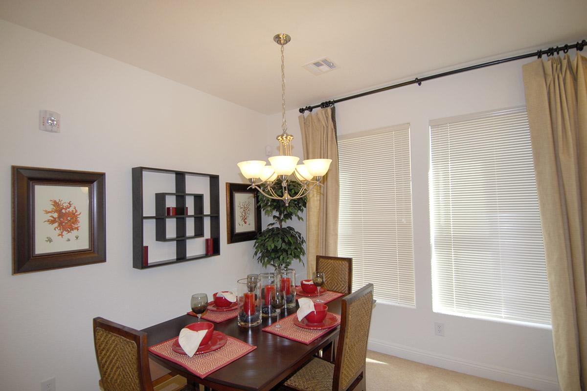 A cozy dining area featuring a wooden table set with red and white tableware, surrounded by two wicker chairs. A decorative chandelier hangs above the table. Walls are adorned with framed artwork and a shelf, with a potted plant in the corner, while natural light filters through the window covered by light curtains.