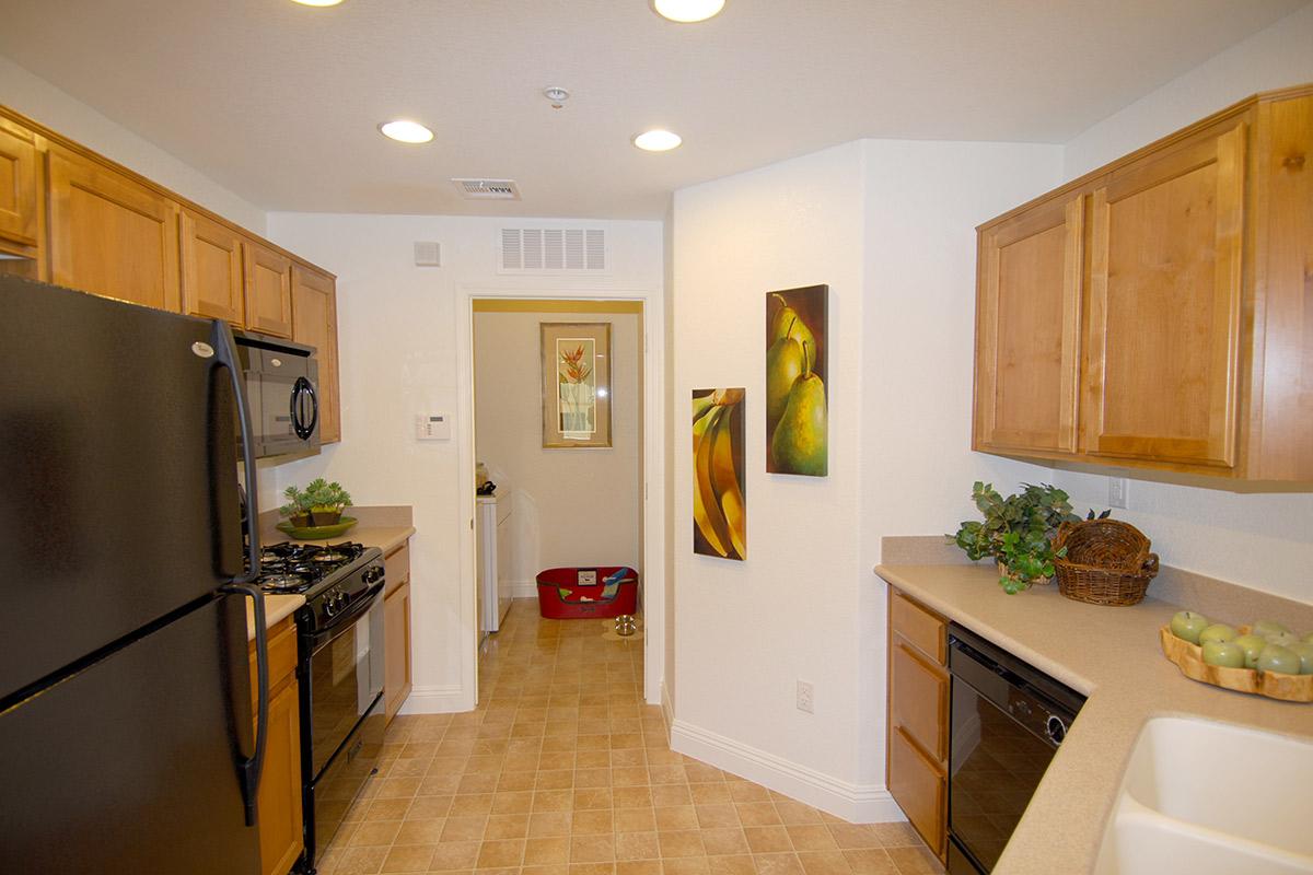 A modern kitchen featuring wooden cabinets, a black refrigerator, and a stove. The countertop is light-colored with decorative items, including a basket with apples. Wall art depicting fruits is displayed, and a doorway leads to a laundry area in the background. Ample lighting illuminates the space.