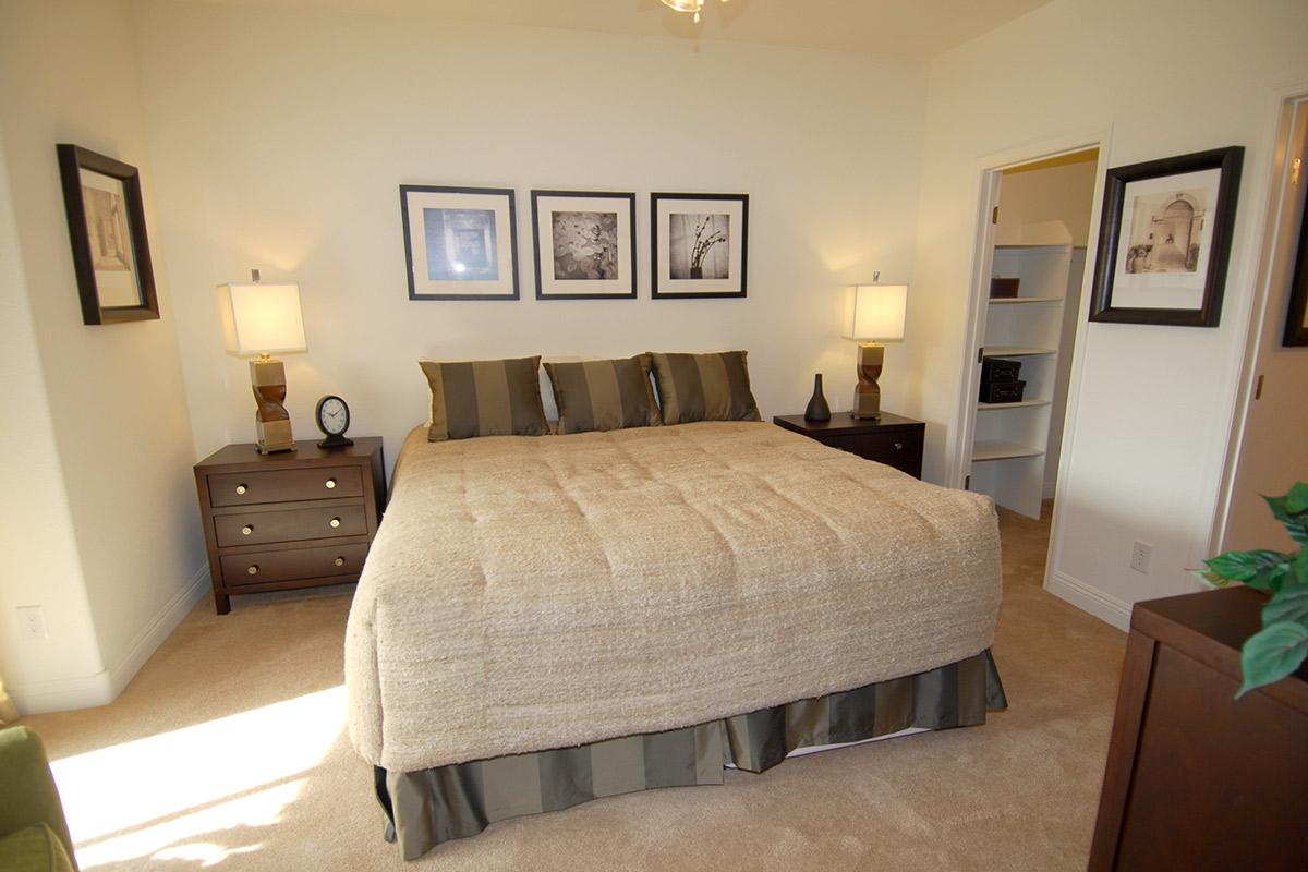 A cozy bedroom featuring a plush bed with a beige coverlet, flanked by two wooden nightstands with lamps. The walls are adorned with framed black-and-white photographs, and a small clock is placed on one nightstand. A green plant adds a touch of color, and there is a glimpse of an open closet.
