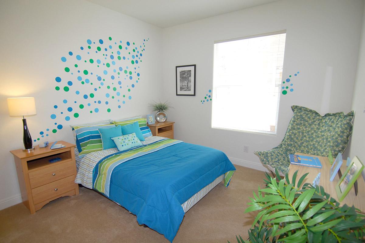 A cozy bedroom featuring a blue and green color scheme. The bed is adorned with a striped duvet and decorative pillows. There are two wooden bedside tables, a lamp, and a green patterned chair in the corner. The wall has playful blue and green dot decorations, and a window allows natural light to enter.