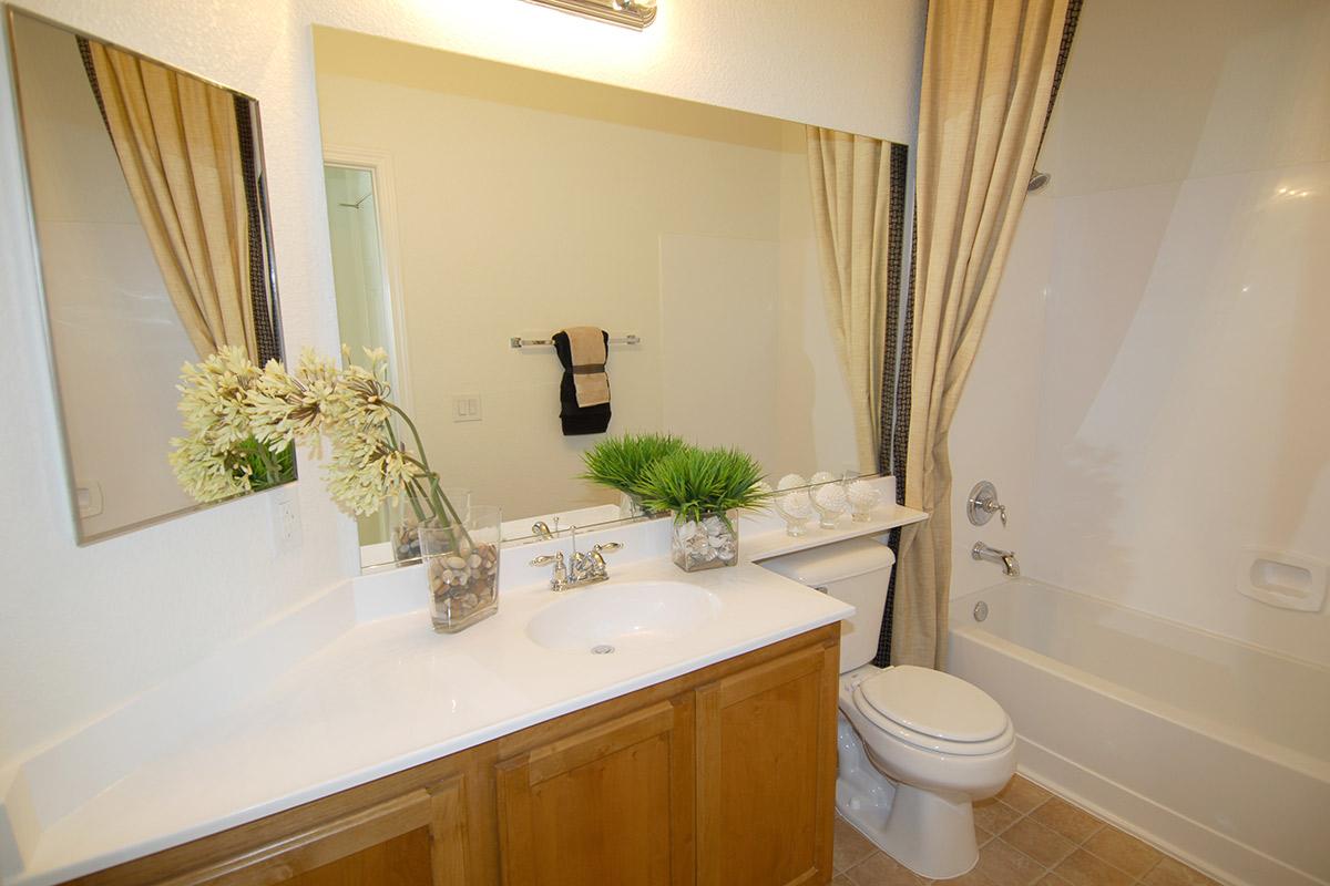 A modern bathroom featuring a white countertop with a sink, a large mirror, and wooden cabinetry. A vase with flowers and a small plant add decor. There is a toilet and a bathtub against the wall, with beige tiles on the floor and a towel hanging on a rack. Natural light brightens the space.