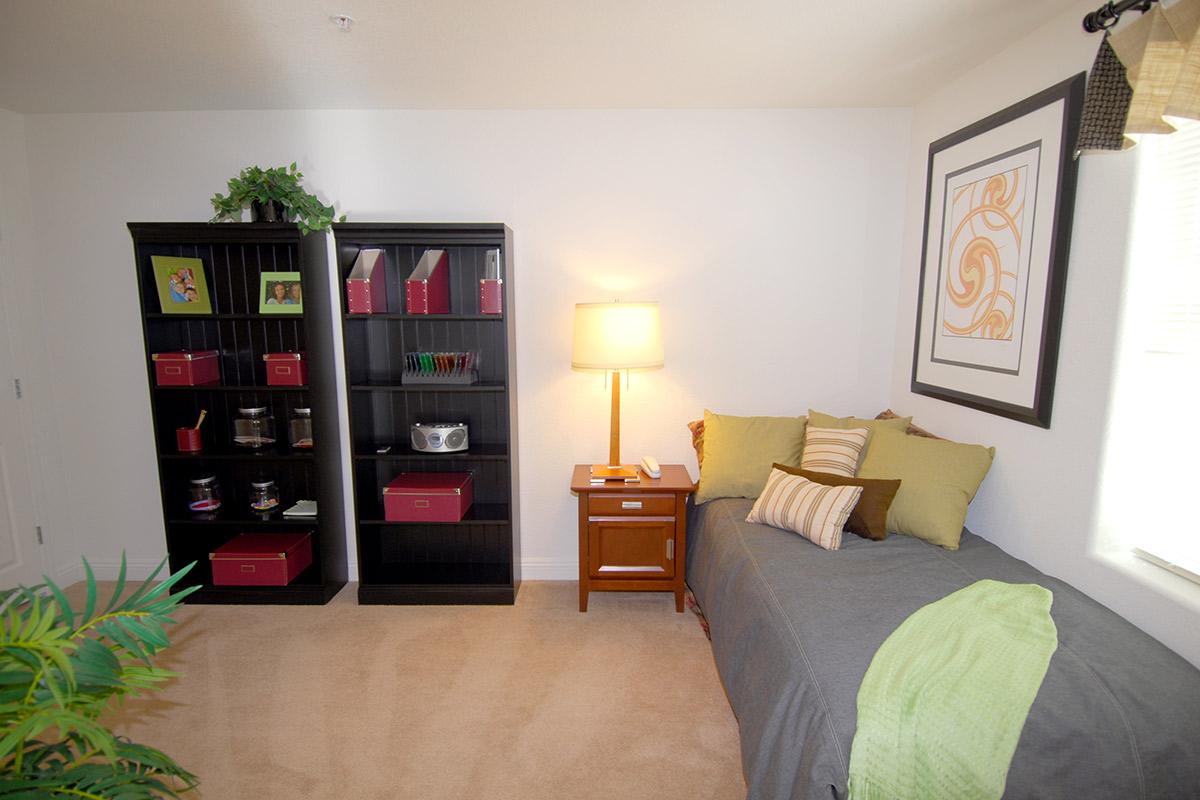 A cozy room featuring a single bed with green and brown accent pillows, a bedside table with a lamp, and two black bookshelves filled with red boxes and decorative items. A potted plant adds a touch of greenery, and a framed artwork in warm tones is mounted on the wall. Natural light filters through a window.