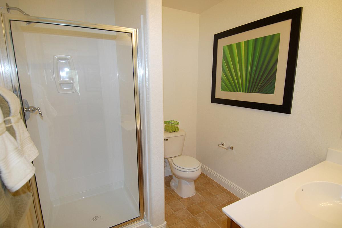 A clean, modern bathroom featuring a glass shower stall, white toilet, and a sink. The walls are painted light colors, and there is a large framed green leaf print hanging on the wall. Soft towels are neatly hung, and the floor is tiled with a light brown pattern.