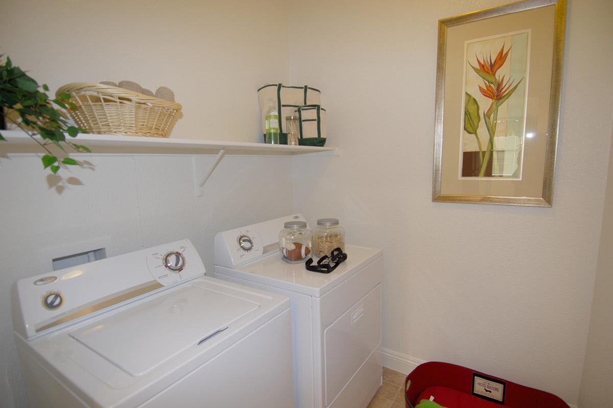 A laundry room with a washing machine and dryer side by side. Above them is a shelf with a basket and decorative jars. A framed artwork depicting a flower hangs on the wall, and a red laundry basket is visible on the floor. The walls are painted a light color, creating a bright and airy atmosphere.