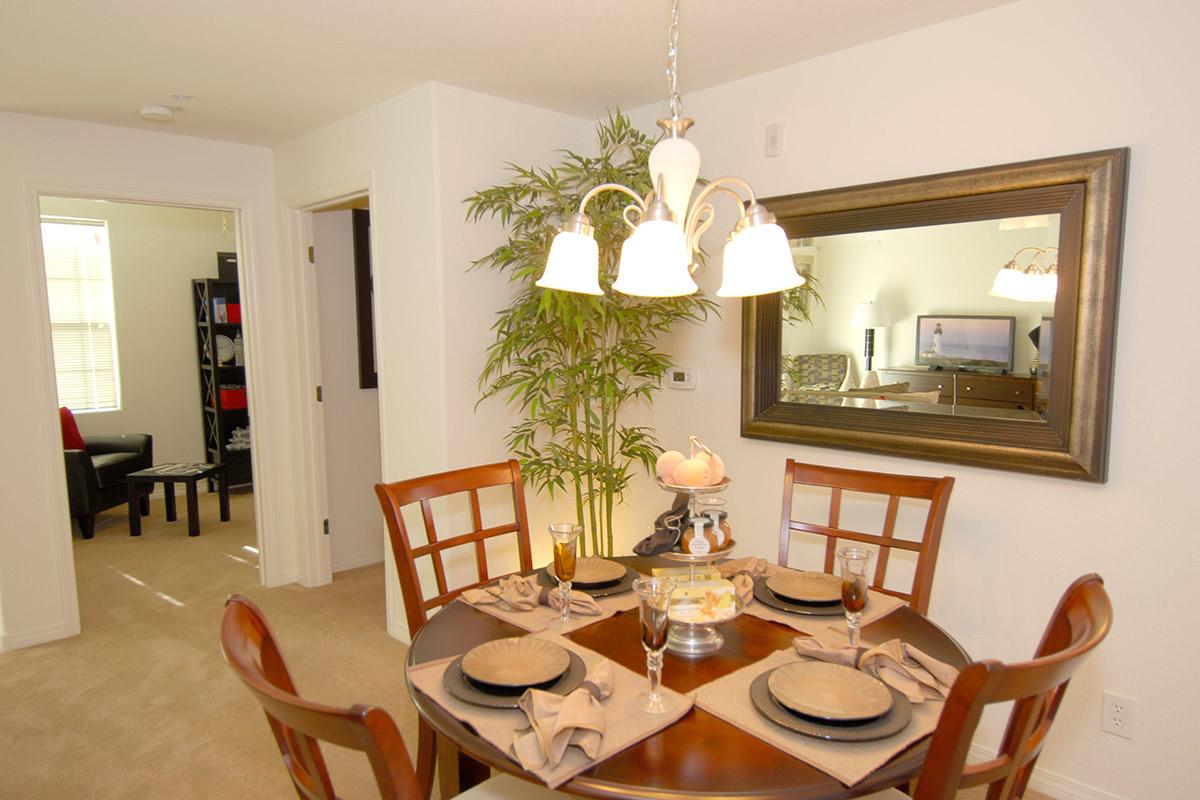 A cozy dining area featuring a round table set for four with place settings, a decorative centerpiece, and a chandelier overhead. The room is bright, with a mirror reflecting a living space, while a potted plant adds a touch of greenery. Soft carpet and neutral-colored walls create an inviting atmosphere.