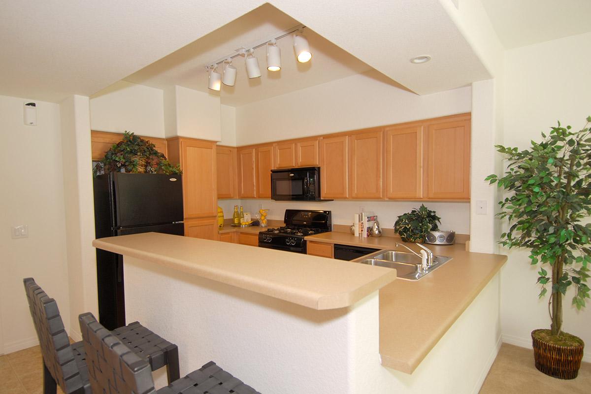 Modern kitchen featuring light wood cabinetry, a black refrigerator, and a stove with an oven. There is an island with bar stools, a sink, and a microwave. Decorative plants add a touch of greenery to the space, illuminated by recessed lighting. The overall design is open and inviting.